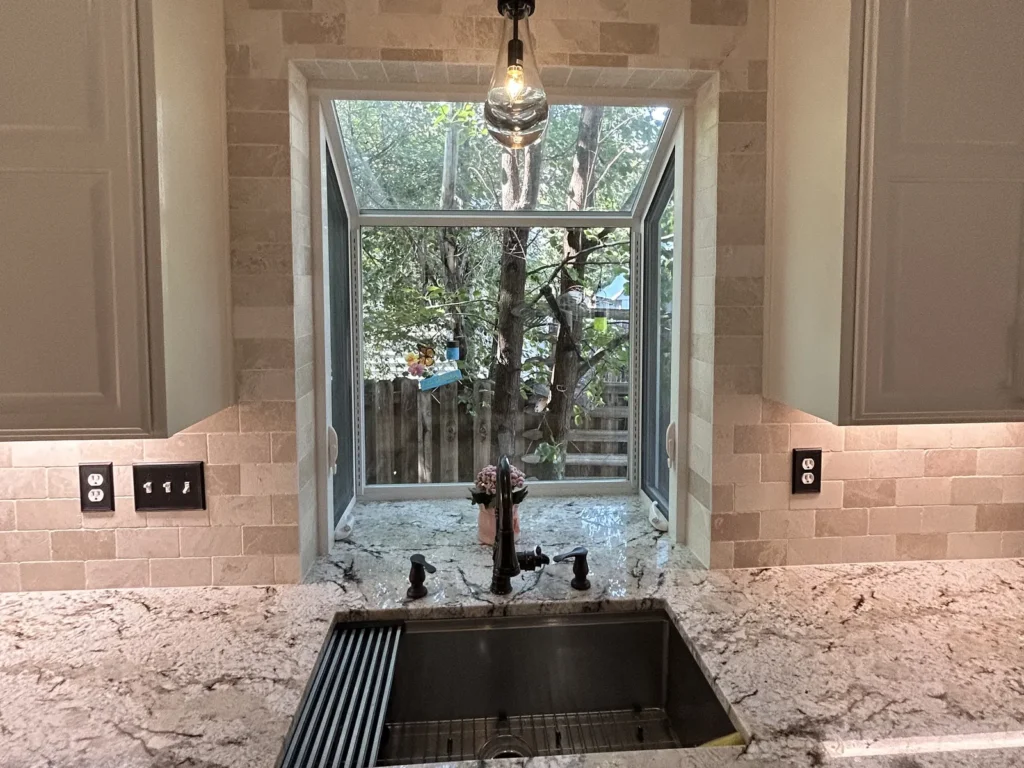 Garden window over a stainless workstation sink with a stone tile surround and pendant light
