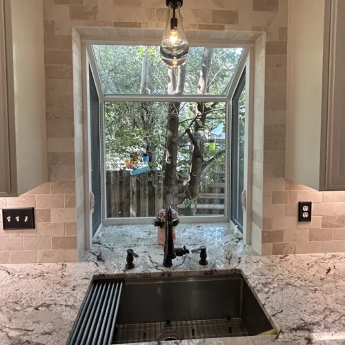 Garden window over a stainless workstation sink with a stone tile surround and pendant light